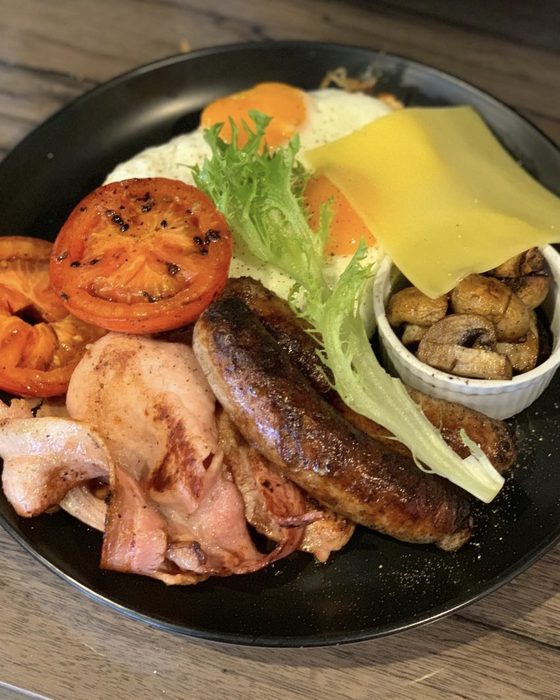 A big breakfast plate with fried eggs, bacon, sausage, grilled tomato, cheese and a pot of mushrooms.