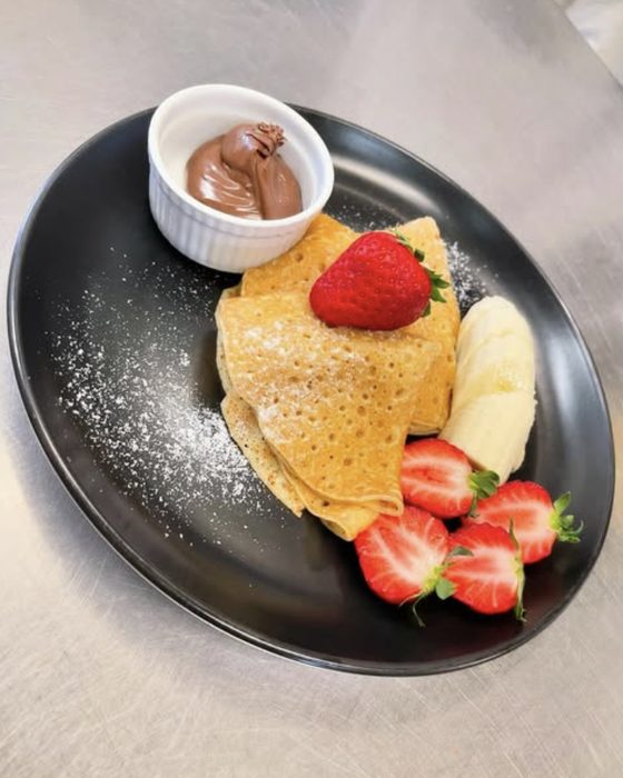 A folded crêpe dusted with icing sugar, served with strawberries, banana and a pot of Nutella on a black plate.