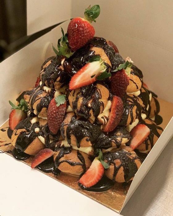 A pyramid of cream-filled profiteroles under dark chocolate sauce and fresh strawberries, in a bakery box.